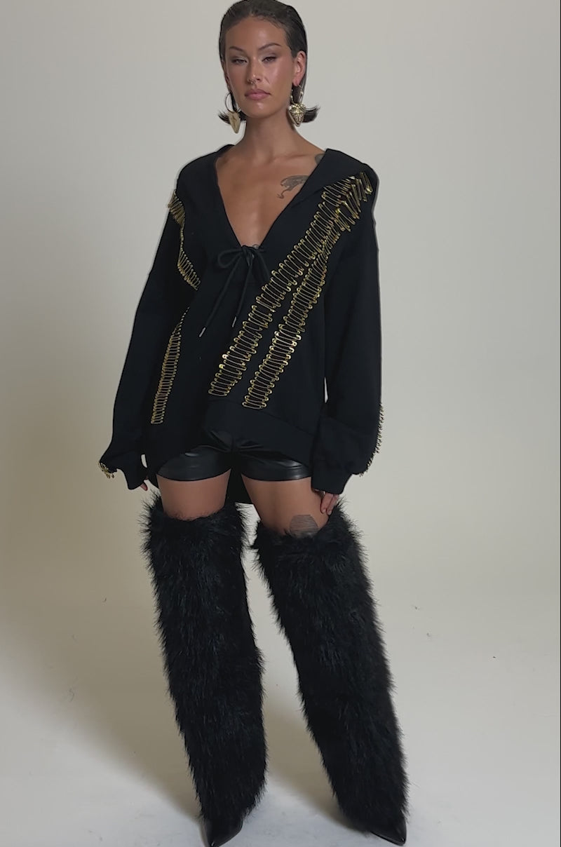 A video of a woman against a plain background in the UNPIN ME OVERSIZED SWEATSHIRT with gold safety pin details, black leather shorts, large hoop earrings, and tall black faux fur boots.