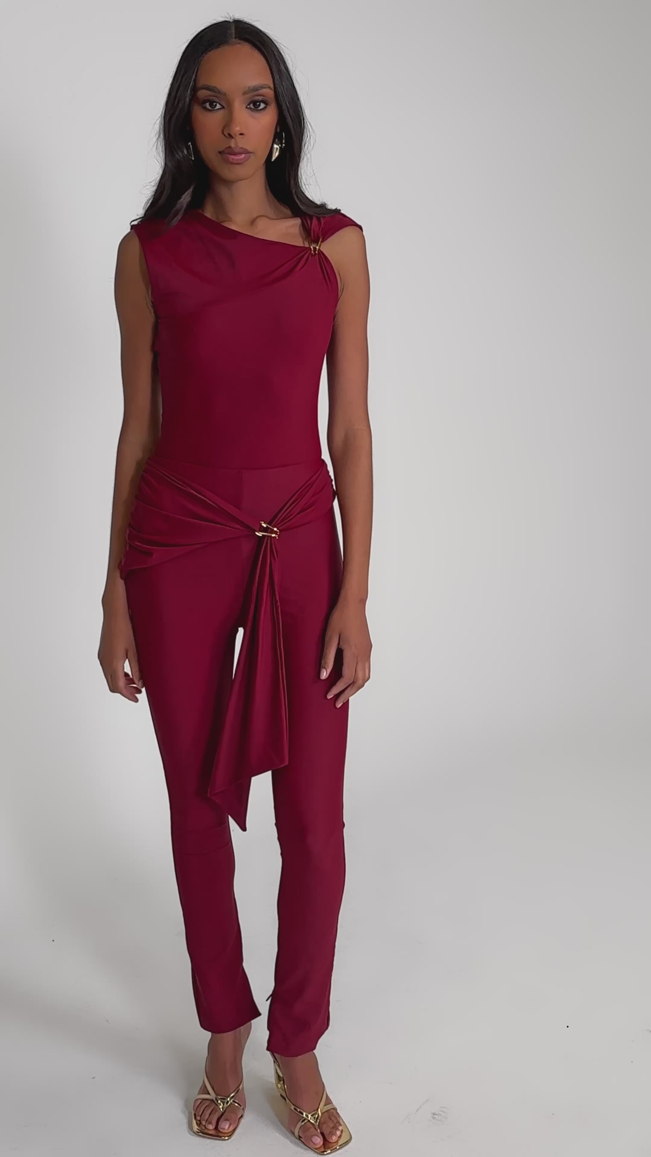 Video of woman modeling the IZZY SEAMLESS LEGGING IN BURGUNDY, paired with gold high-heeled sandals and a sleeveless deep red top with a draped sash featuring gold ring detail, set against a plain light background.