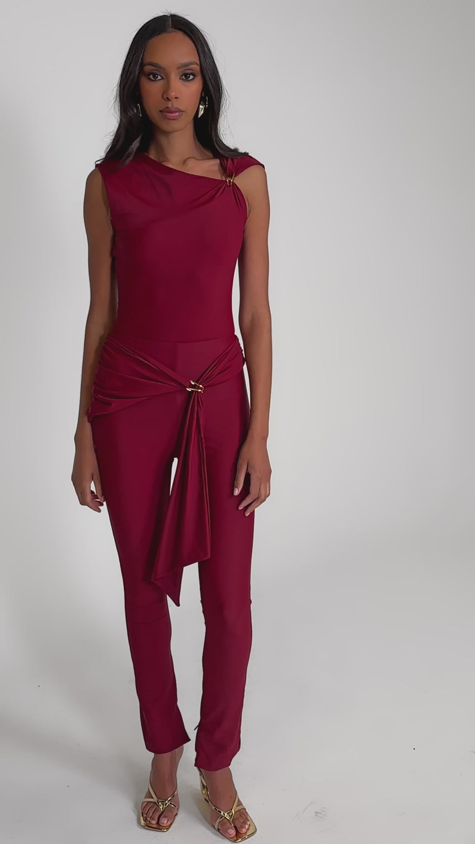 Video of woman modeling the IZZY SEAMLESS LEGGING IN BURGUNDY, paired with gold high-heeled sandals and a sleeveless deep red top with a draped sash featuring gold ring detail, set against a plain light background.