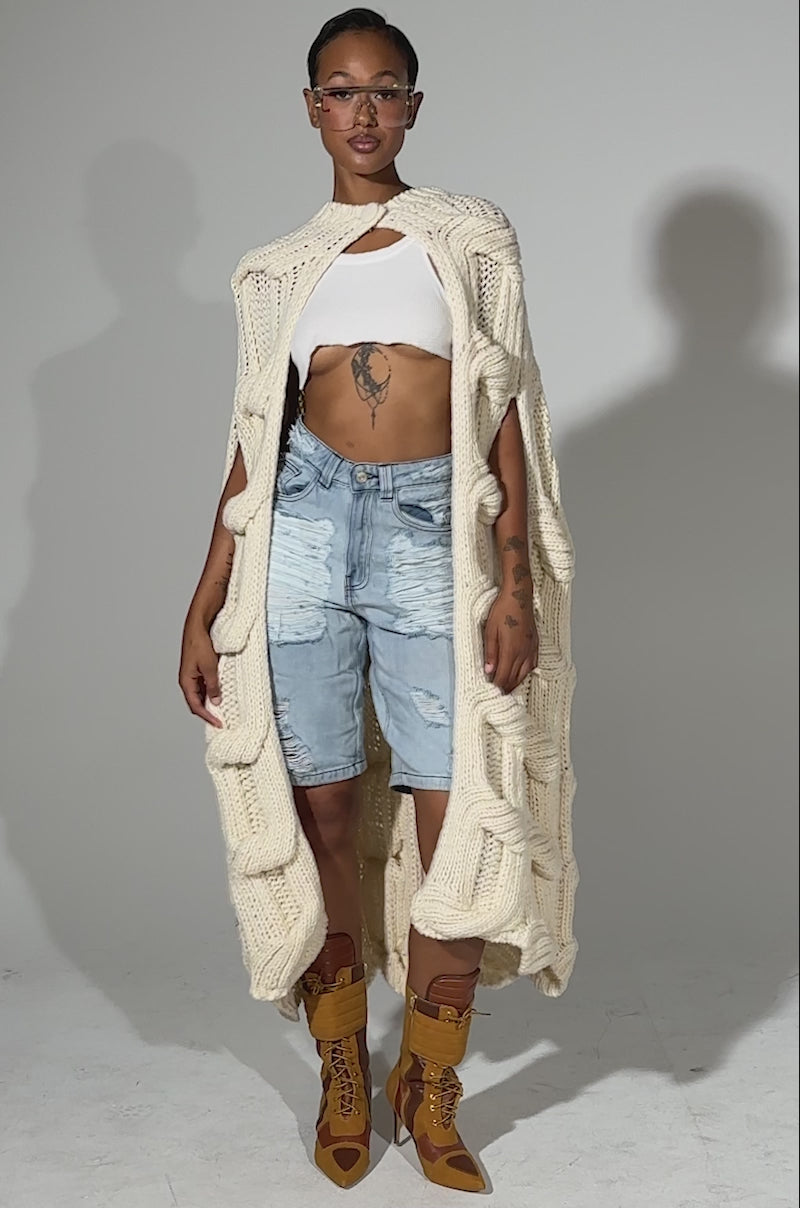 A video of a person in glasses, a white cropped top, light blue high-waisted denim shorts, tan lace-up boots, and the TEXTURED CHUNKY KNIT SWEATER standing against a plain light background.