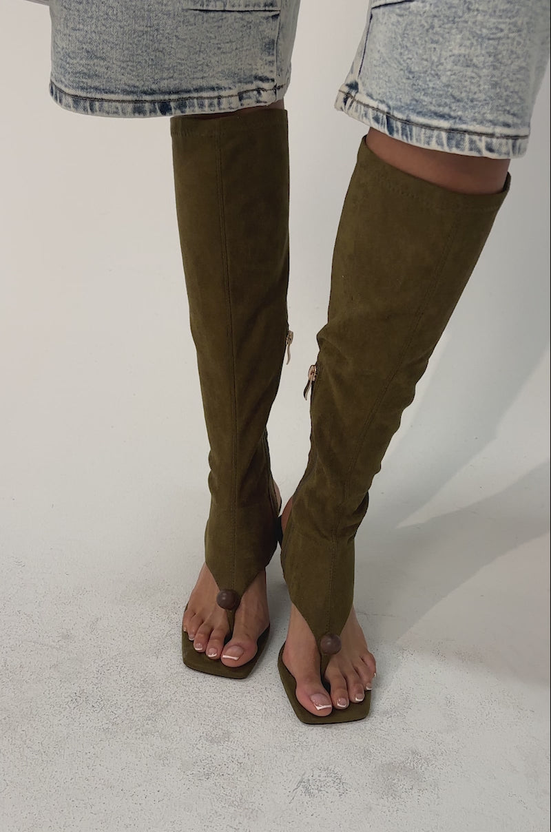 Video of woman modeling the AZALEA WANG DOTTIE Olive Stretch Faux Suede Boot with embellished heel and toe hardware, knee-high shaft, side zippers, and holding a textured brown handbag.