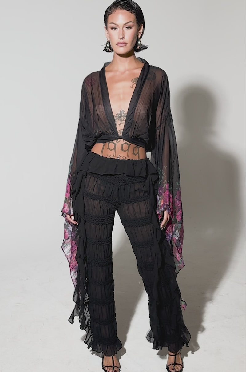 Video of woman modeling the DANCING QUEEN RUFFLE PANT, featuring a high-rise fit, sheer black fabric, and horizontal ruched bands, styled with open-toe heels. 