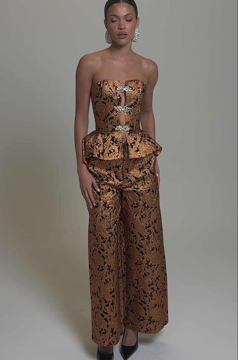 Video of woman modeling the AWAY FROM ME BROCADE CORSET, a brown and gold peplum top with ornate patterns and jeweled clasps, styled with matching fitted pants and statement earrings against a neutral background.