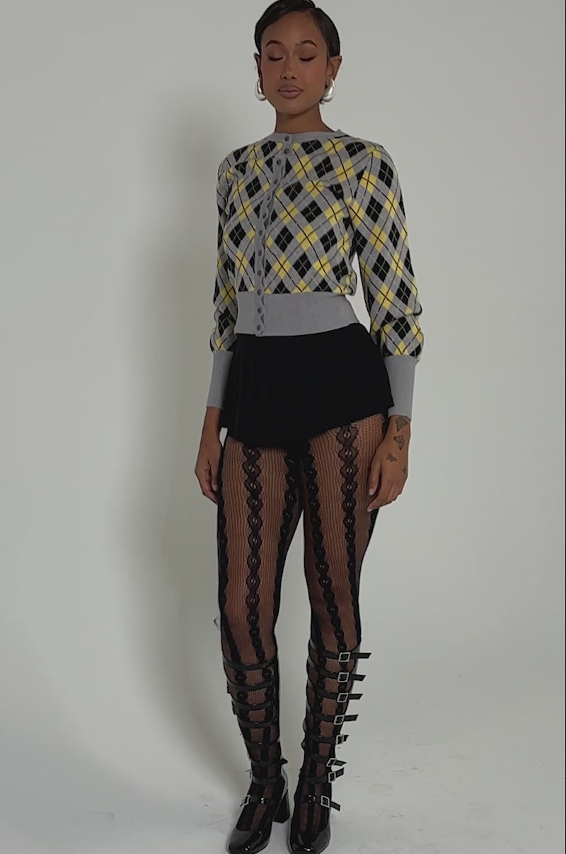 Video of woman modeling the GUESS WHAT PLAID CARDIGAN in yellow, gray, and black with gray trim, a black pleated mini skirt, and black lace tights stands against a light background, confidently facing the camera.