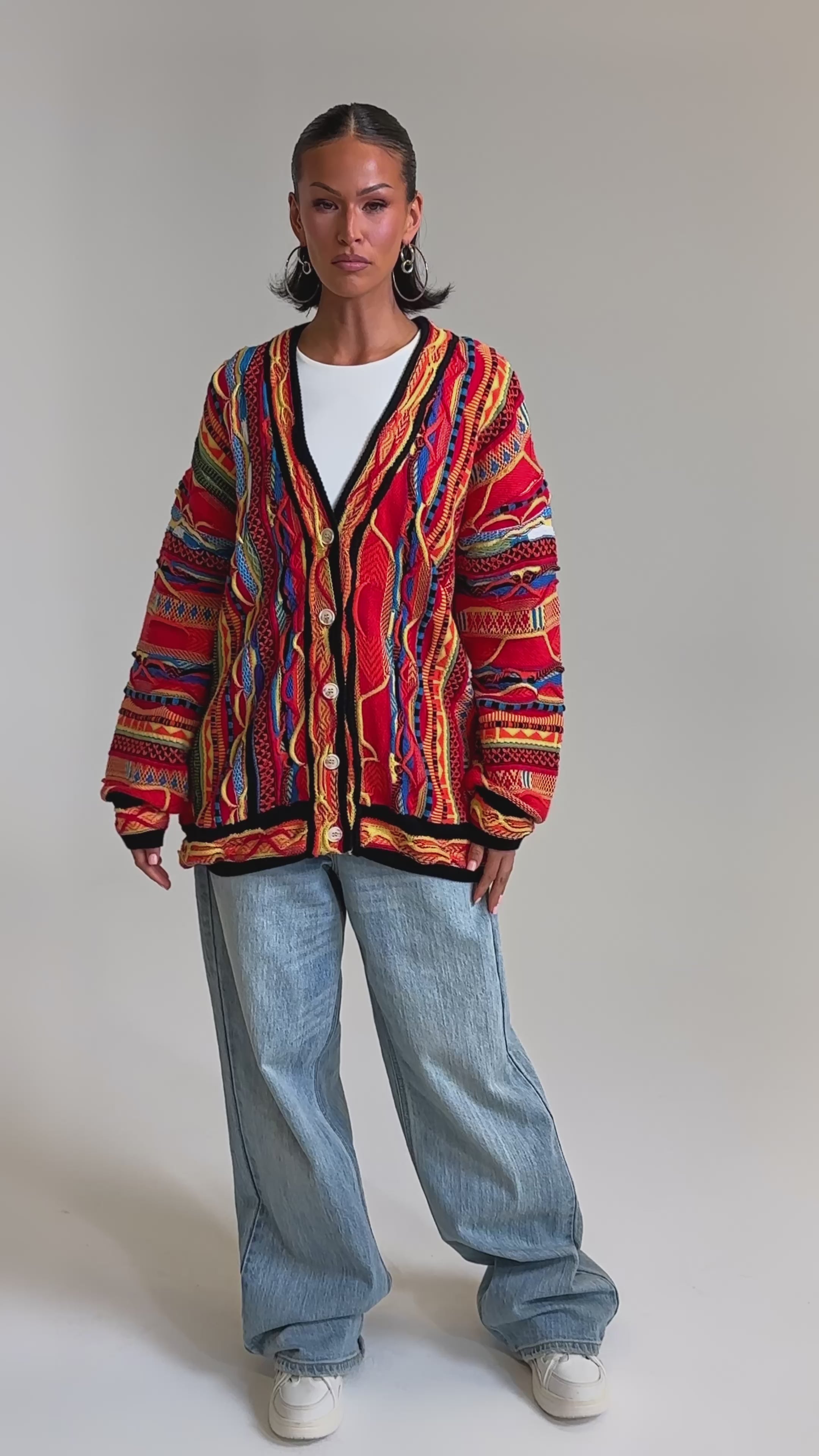 A video of a woman wearing the CRAZY IN LOVE CARDIGAN, a colorful retro knit, over a white shirt with wide-leg light blue jeans and white sneakers. She accessorizes with hoop earrings and has two ponytails, set against a plain light background.
