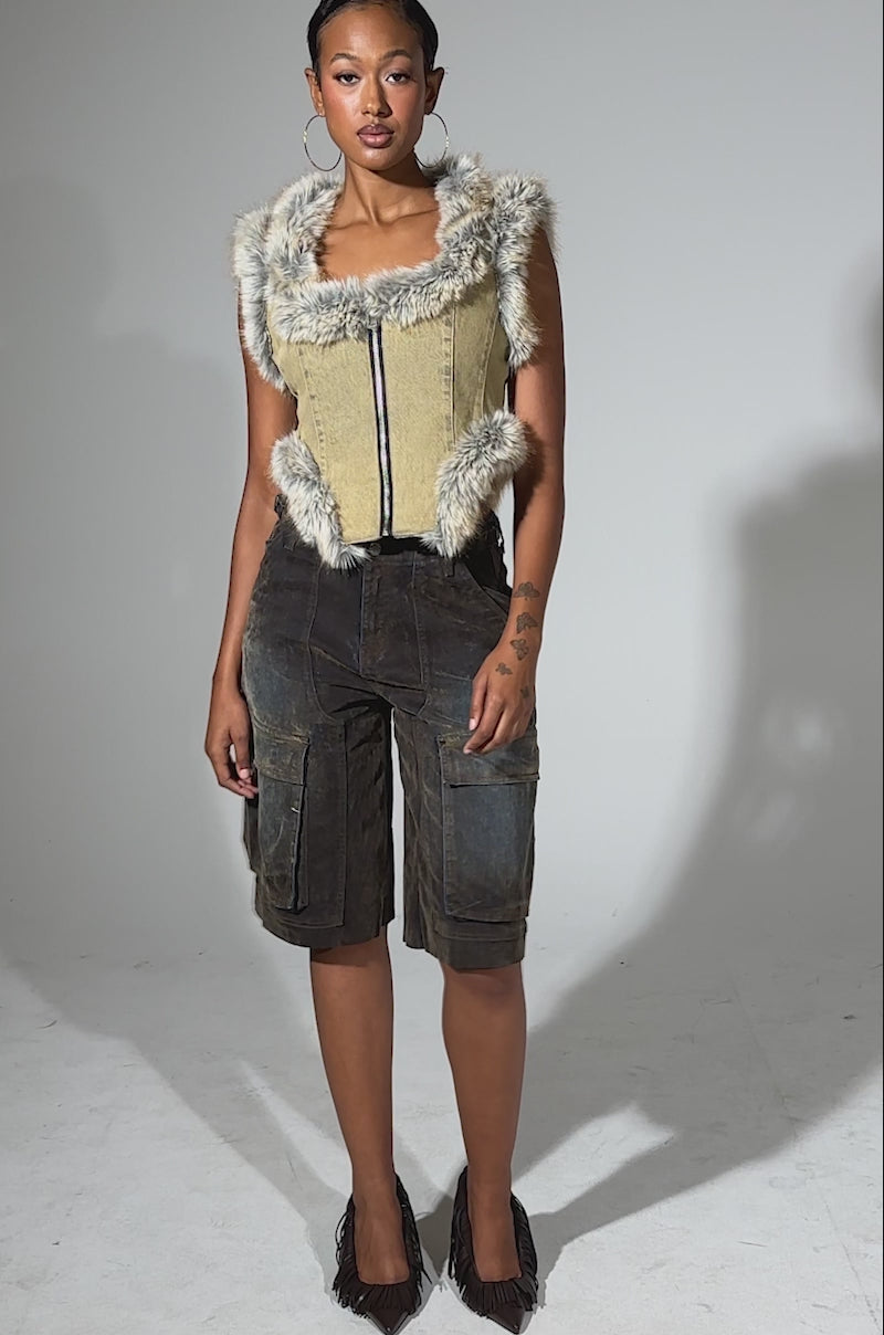 A video of a woman exuding confidence in the ITTY BITTY FUR TRIM VEST with a corset-inspired hem, paired with knee-length dark denim shorts, hoop earrings, and black heels. Her hair is pulled back against a plain light gray background.