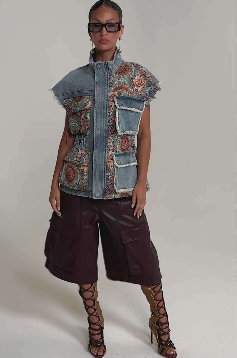A video of a woman in the KAI PATCHWORK DENIM VEST with colorful embroidery and pockets, brown wide-leg cargo shorts, and knee-high lace-up sandals against a plain background.