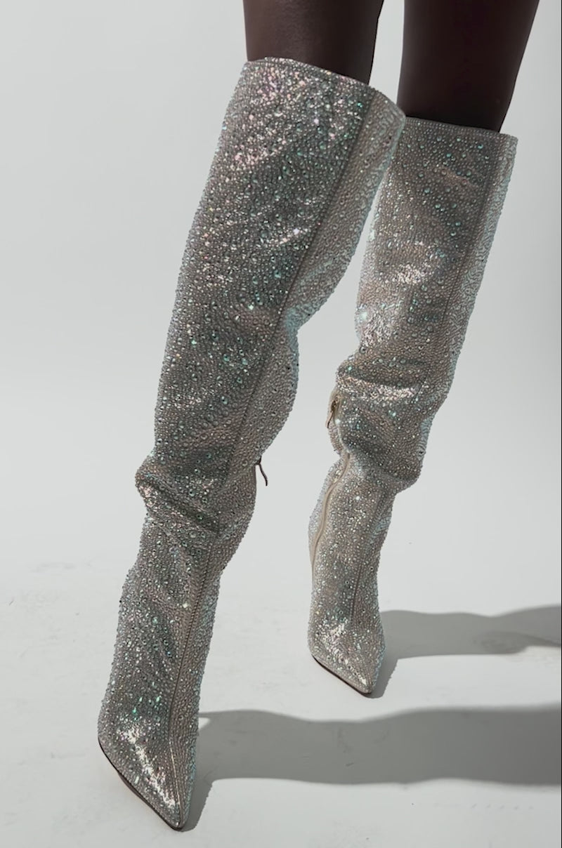 A video of a person wearing the AZALEA WANG BIBBLE Gold Rhinestone Stiletto Over The Knee Boot, featuring pointed toes and a visible side zipper, while standing against a neutral background.