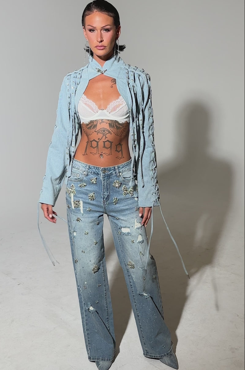 A video of a person in the IMPULSIVE DENIM BOLERO with lace-up sleeves, paired with matching rhinestone-studded jeans and a white lace bralette. Their midriff, featuring a "1997" tattoo, is exposed against a neutral background.