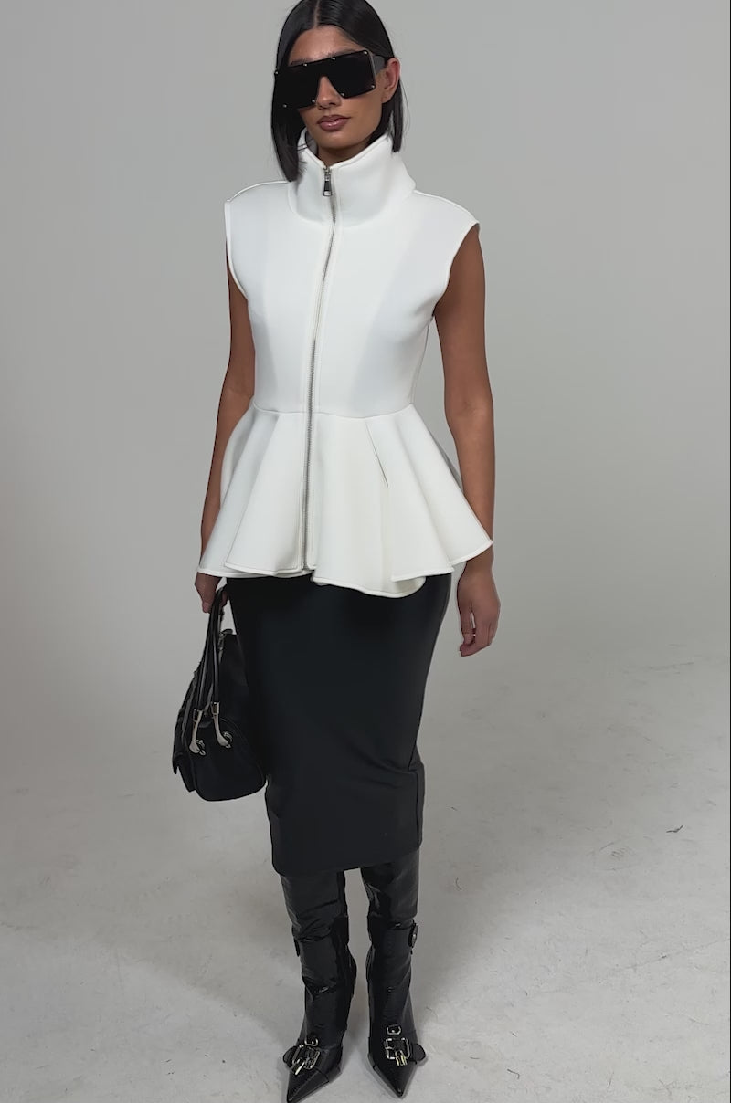 A video of a woman in the LOTTIE NEOPRENE PEPLUM VEST, a fitted black midi skirt, tall black boots, sunglasses, and carrying a black handbag against a plain light background.