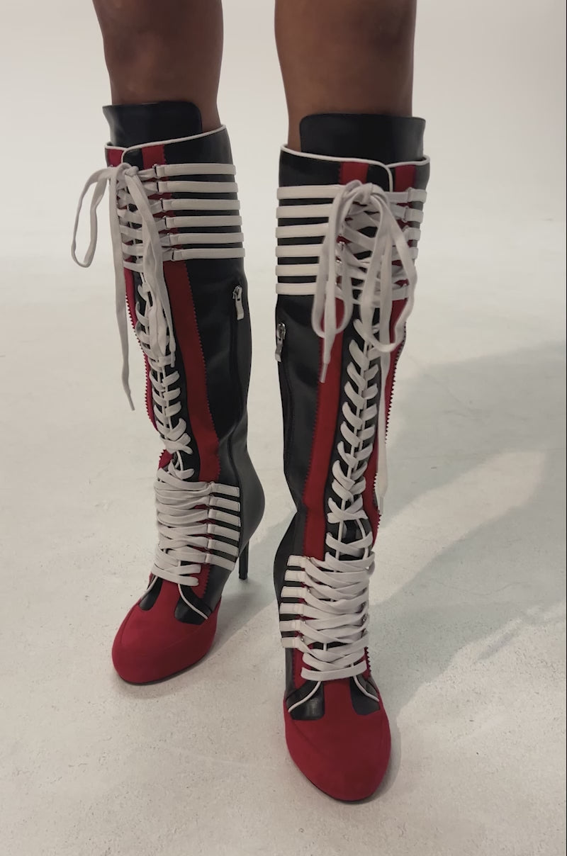 Video of woman modeling the AZALEA WANG NITRO RED MULTI SPORTY STILETTO BOOT—knee-high boots in black, red, and white with color blocking, white lace-up fronts, red accents, stripes, and side zippers. Their legs are visible from the knees down.