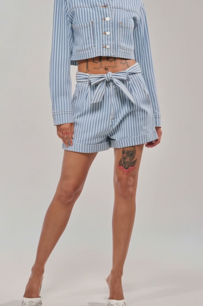 Video of woman modeling the VACA VIBES SHORT, a blue and white nautical striped high-rise short with a tie belt, paired with a matching cropped jacket, white heels and hoop earrings
