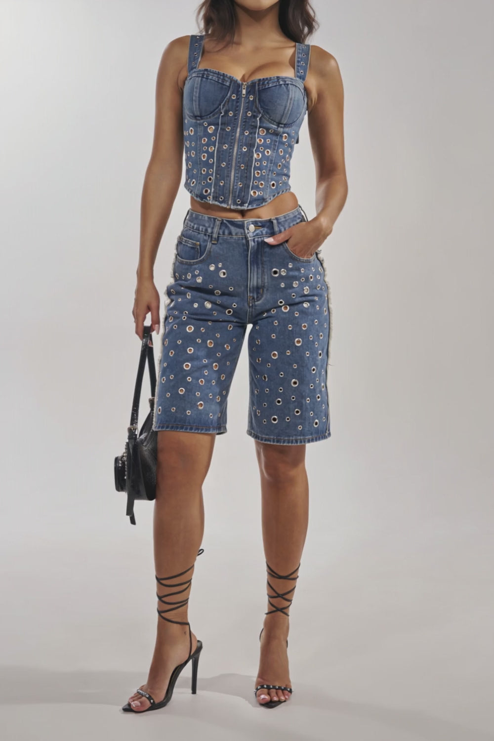 Video of woman modeling the TAYLOR DENIM BERMUDA WITH GROMMET—high-rise fit shorts featuring silver grommet hardware and a frayed hem—paired with strappy black high-heeled sandals.