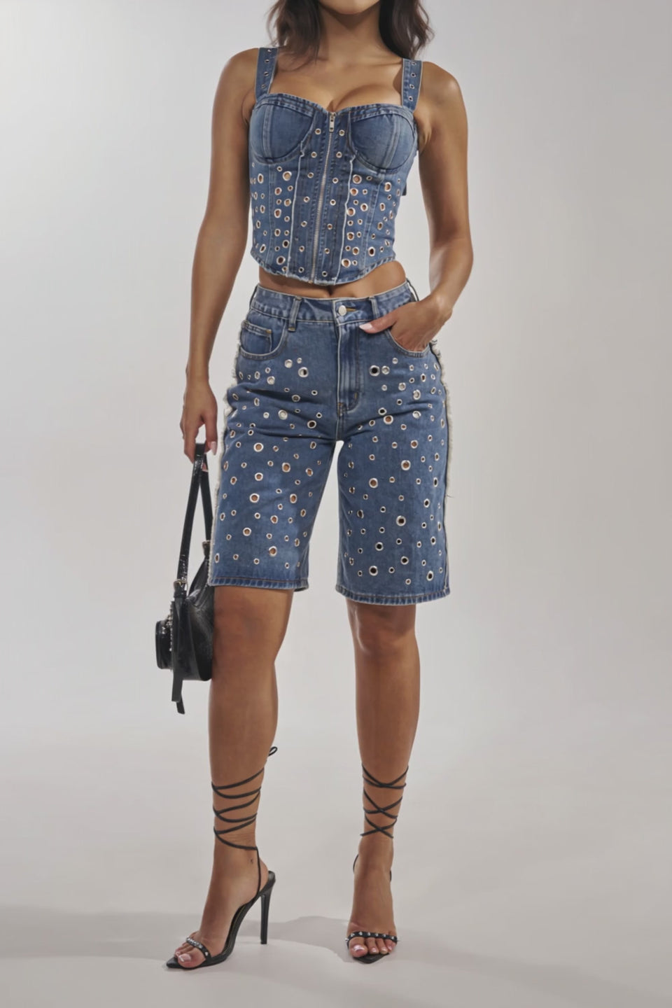 Video of woman modeling the TAYLOR DENIM BERMUDA WITH GROMMET—high-rise fit shorts featuring silver grommet hardware and a frayed hem—paired with strappy black high-heeled sandals.