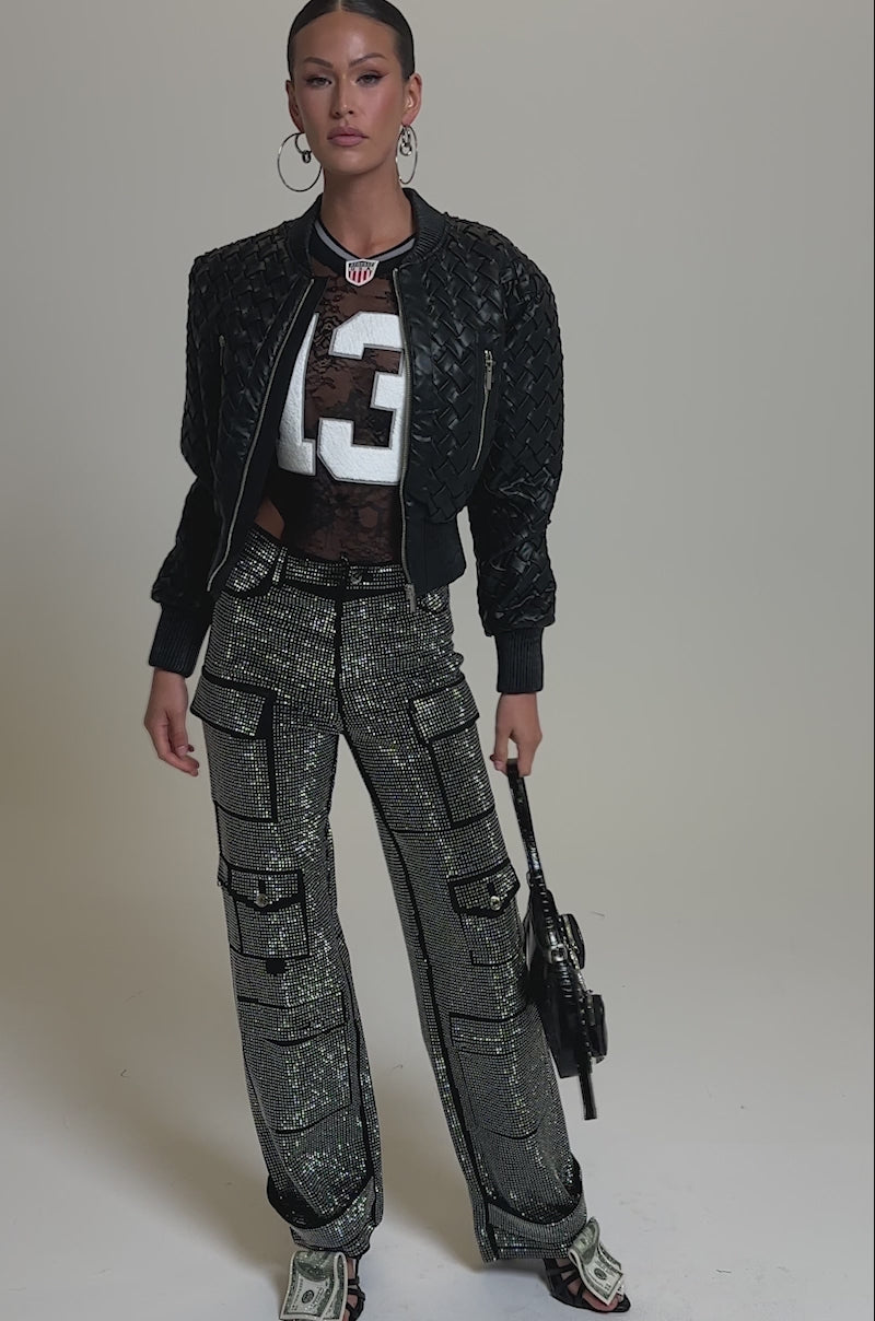 A video of a woman in AMMO BONNE SOIREE RHINESTONE CARGO PANTS IN BLACK, a black textured jacket, sheer top with "13", hoop earrings, a small handbag, and open-toe heeled boots against a plain background.