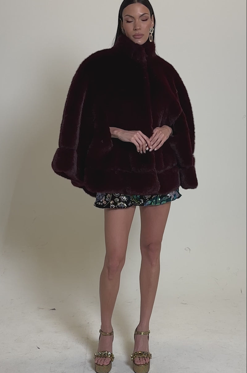 A video of a woman wearing the CELINE FAUX FUR CAPE in deep burgundy, paired with a short floral skirt, gold platform heels, and large gold earrings.