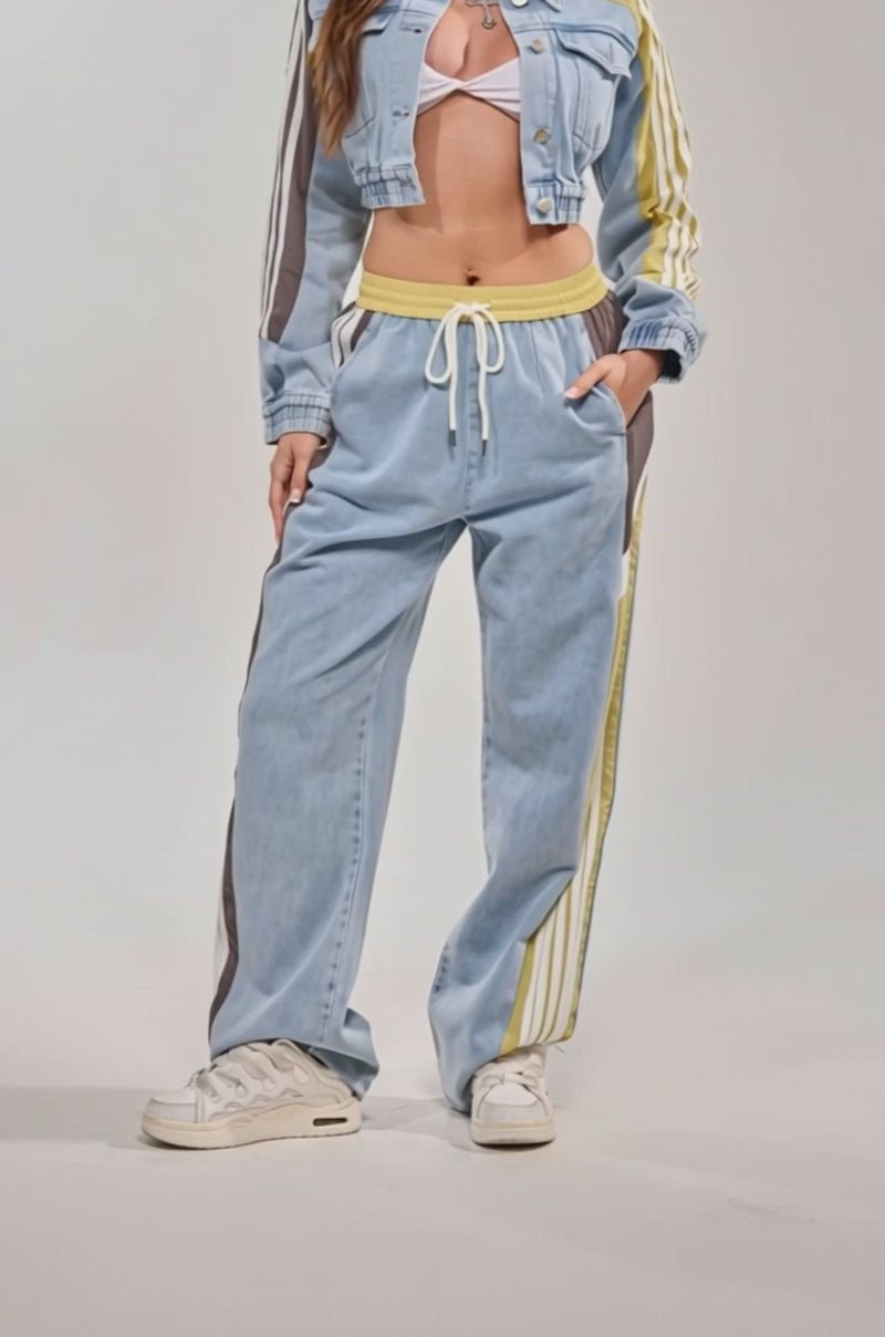 A video of a person wearing ALONE AGAIN DENIM TRACK PANT—light blue joggers with yellow, brown, and white stripes, a high-rise elastic waistband with drawstrings—and white sneakers. 