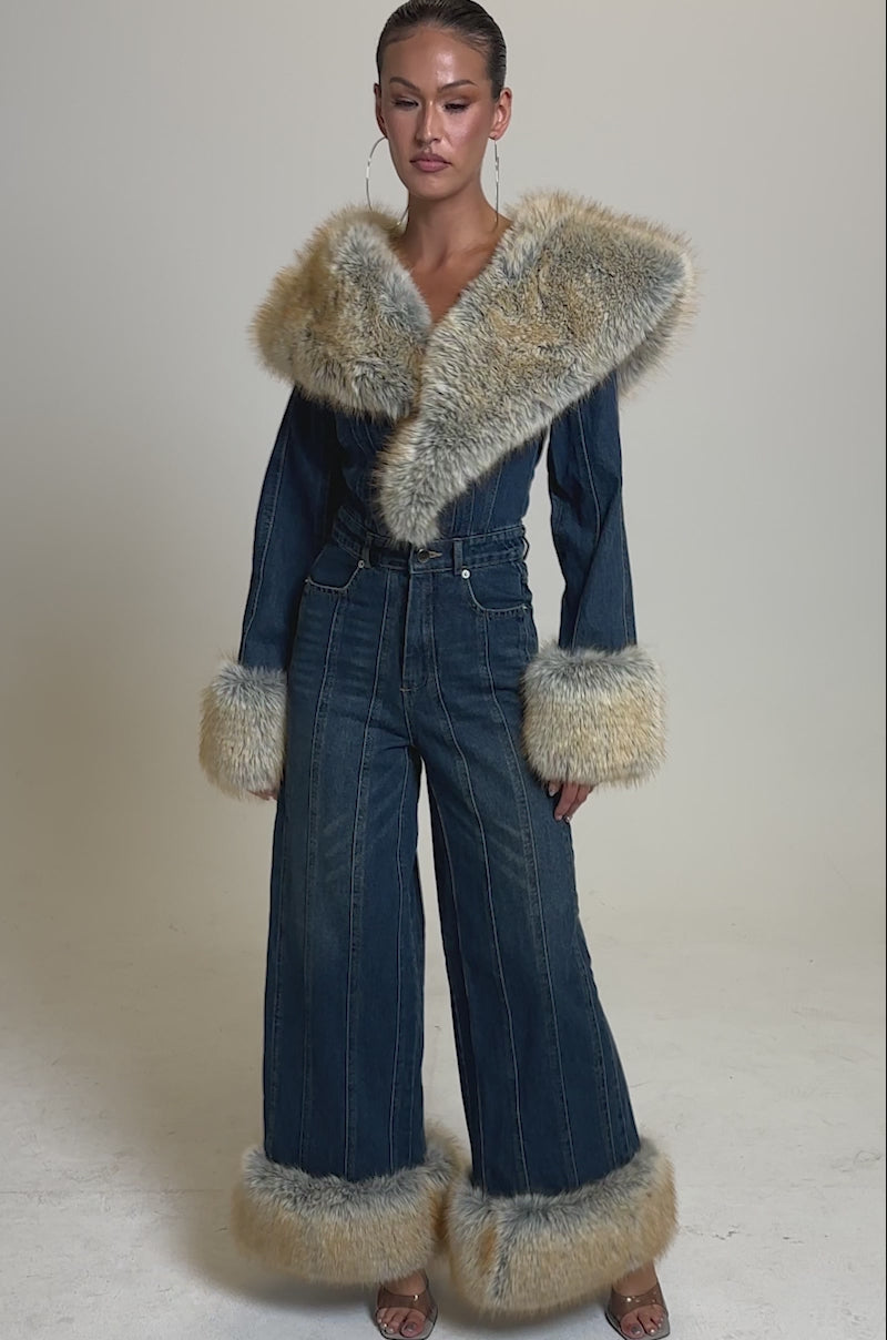 A video of a woman in the LENNOX DENIM JUMPSUIT WITH FUR ACCENTS, featuring wide legs and faux fur details on the collar, cuffs, and hems, styled with slicked-back hair, hoop earrings, and heeled sandals.