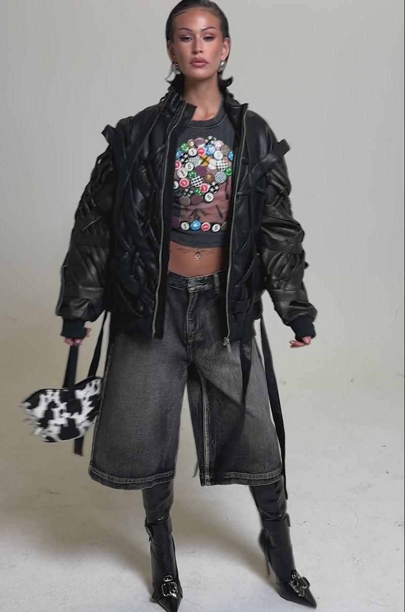 A video of a woman wearing the LOCKED IN STRAP BOMBER, a cropped shirt with colorful buttons, wide-leg faded jeans, tall black boots, and carrying a black-and-white patterned handbag.
