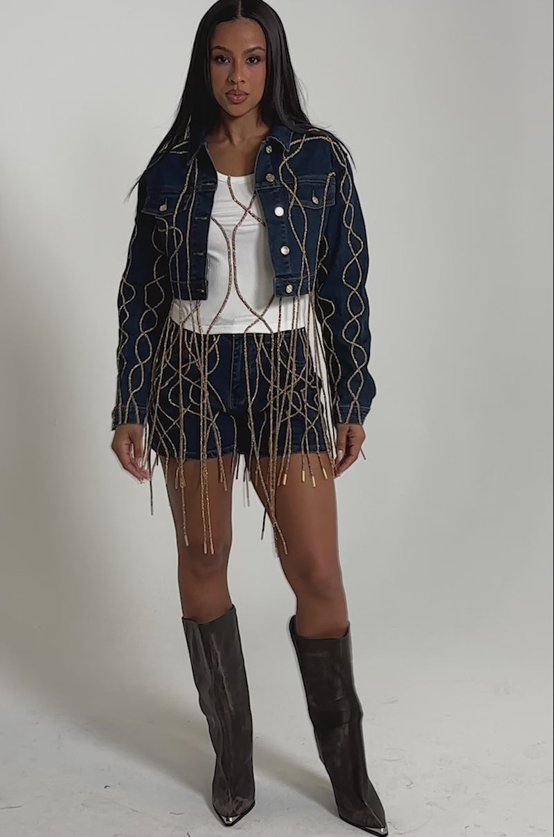 A video of a model wearing the NINA EMBELLISHED RIBBED TANK with decorative gold chains, paired with dark denim shorts and tall gray boots.