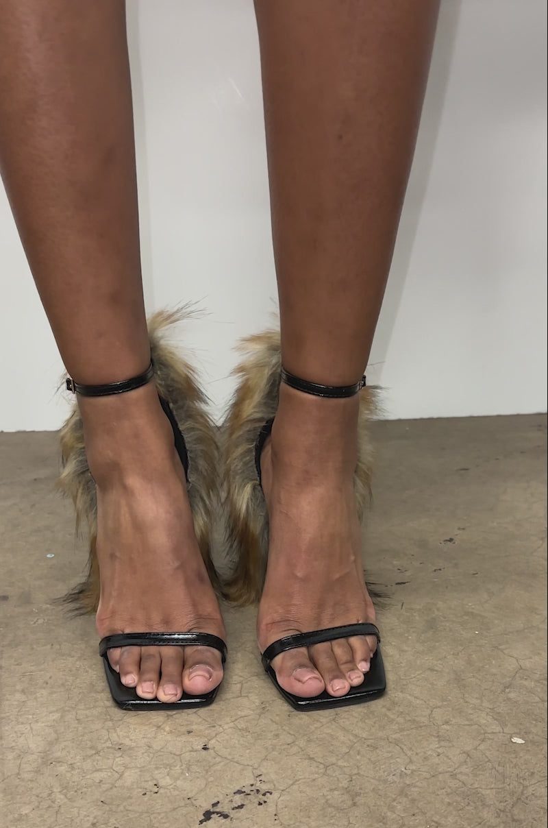 A video of a person wearing the AZALEA WANG ARETTE Black Sandal with Brown Fur standing on a concrete floor against a white wall.