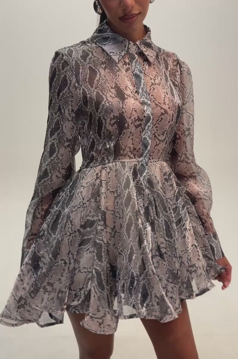 Video of a woman modeling the A woman poses against a plain background wearing the WILD RHYTHM SNAKE PRINT MINI DRESS, a long-sleeve, collared mini dress with rhinestone embellishments, sheer snake-print fabric, and a fit-and-flare silhouette featuring a flared skirt.
