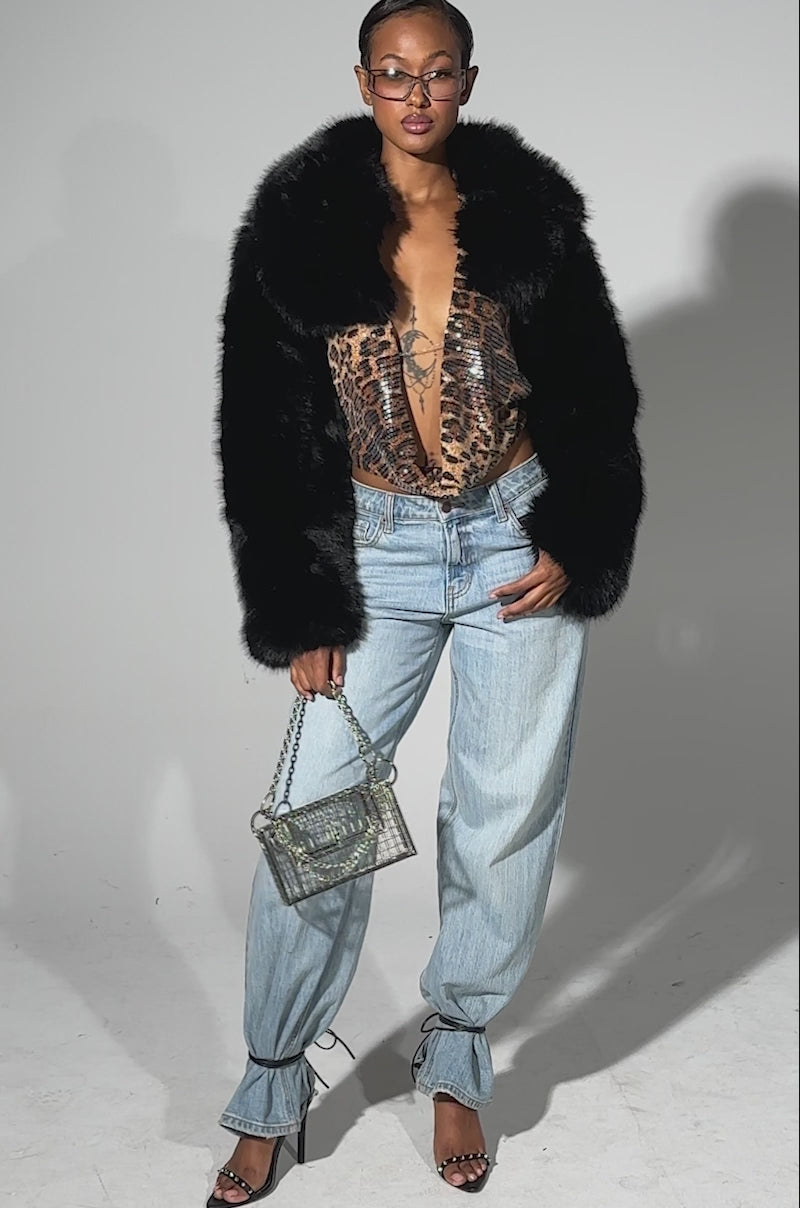 A video of a woman wearing the LEILA ULTRA CROP FAUX FUR IN BLACK, paired with a patterned top, light blue ankle-tie jeans, heeled sandals, a small metal cage handbag, and glasses, standing before a plain light background.