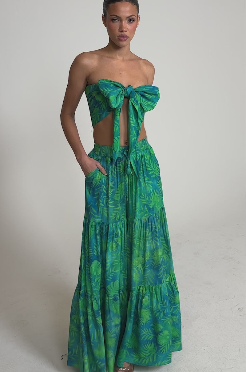 Video of woman modeling the INTO THE GARDEN BRALETTE IN GREEN, a green and blue tropical print top tied in a bow, paired with a matching skirt, standing with hands in pockets against a plain light background.