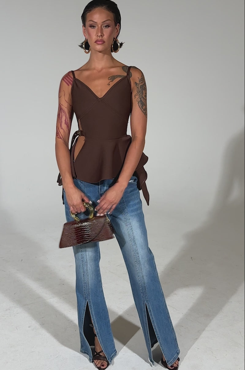 A video of a woman in the TRACY SIDE ADJUSTER PEPLUM TOP, blue wide-leg jeans with front slits, black heeled sandals, large hoop earrings, and a small brown handbag stands against a neutral background.