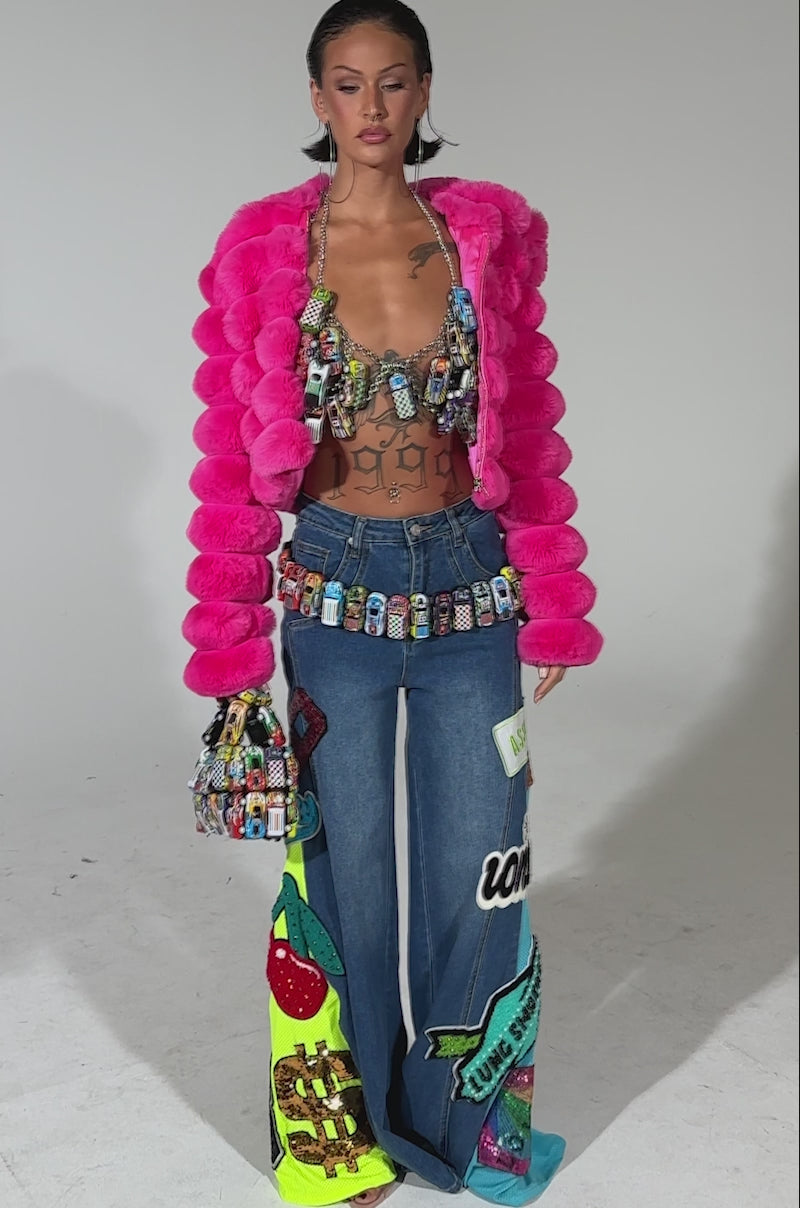 A video of a woman posing in the POPCORN FAUX FUR IN PINK jacket, paired with a beaded bra, wide-leg jeans with colorful appliqué, a jeweled belt, and a patterned handbag against a neutral background.