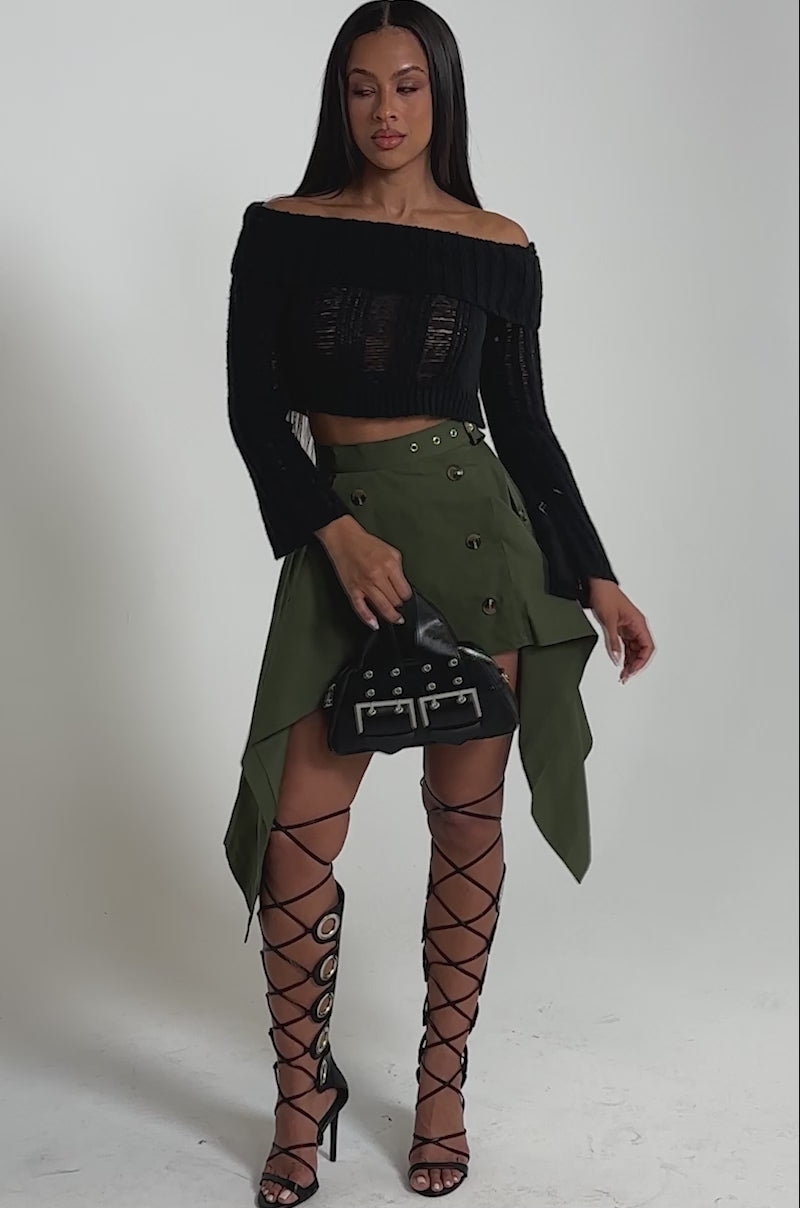 A video of a woman wearing a black off-shoulder cropped sweater, the MAEVE MINI SKIRT IN OLIVE with a handkerchief hem and buttons, tall black lace-up sandals, and holding a small black handbag by her side.