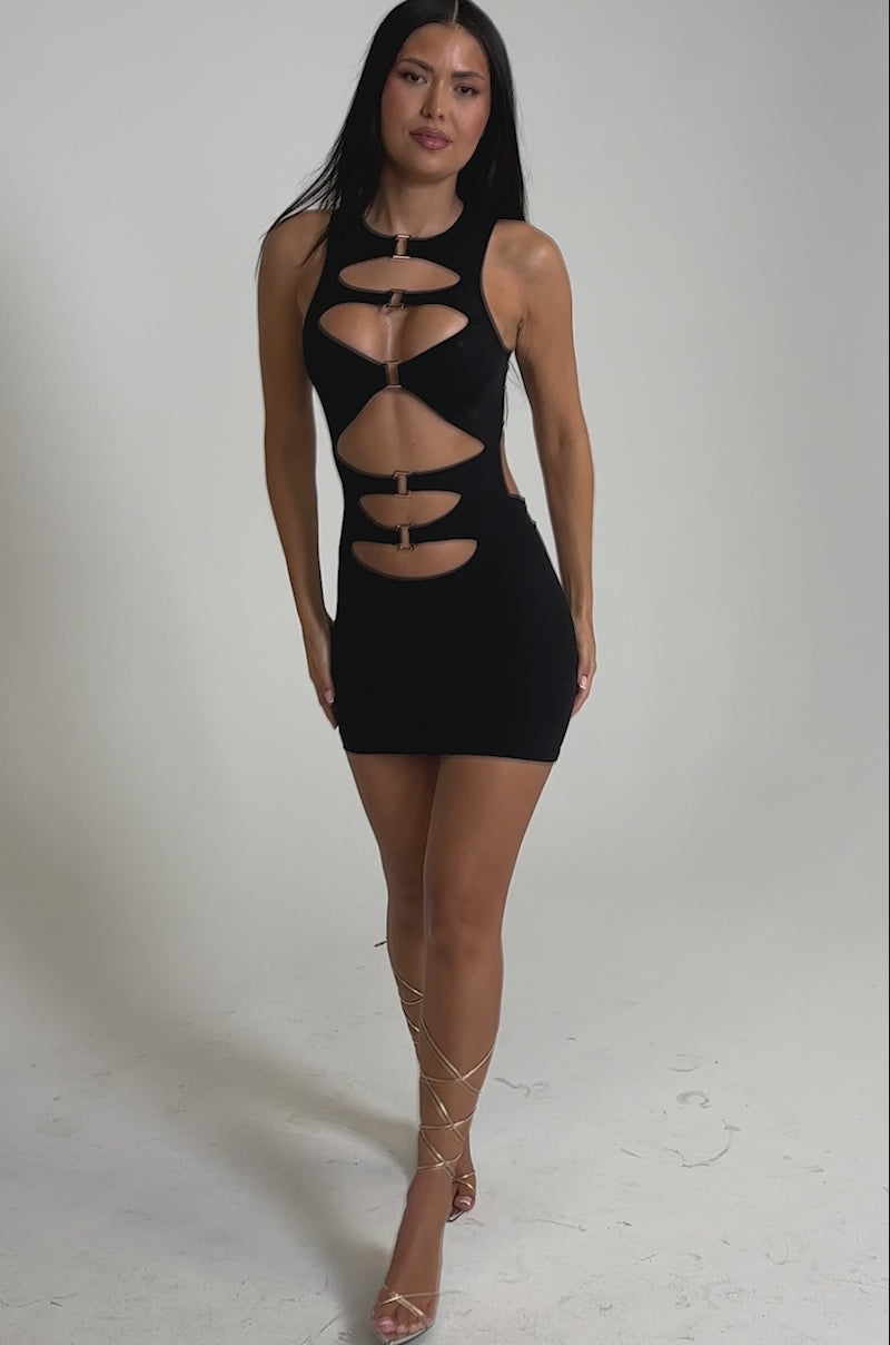 A video of a woman wearing the SIERRA Q MINI DRESS featuring chic cutout mesh with gold accents, paired with clear high heels and lace-up straps around her legs.