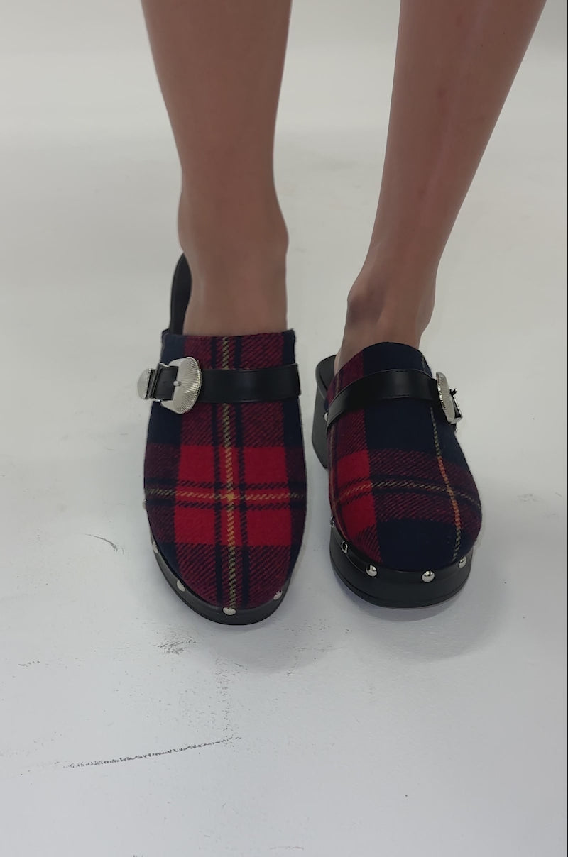 Video of woman modeling the AZALEA WANG LATIFAH RED PLAID HARDWARE FLAT MULE clogs with black straps, silver buckles, and silver-studded black soles stands on a light-colored surface.