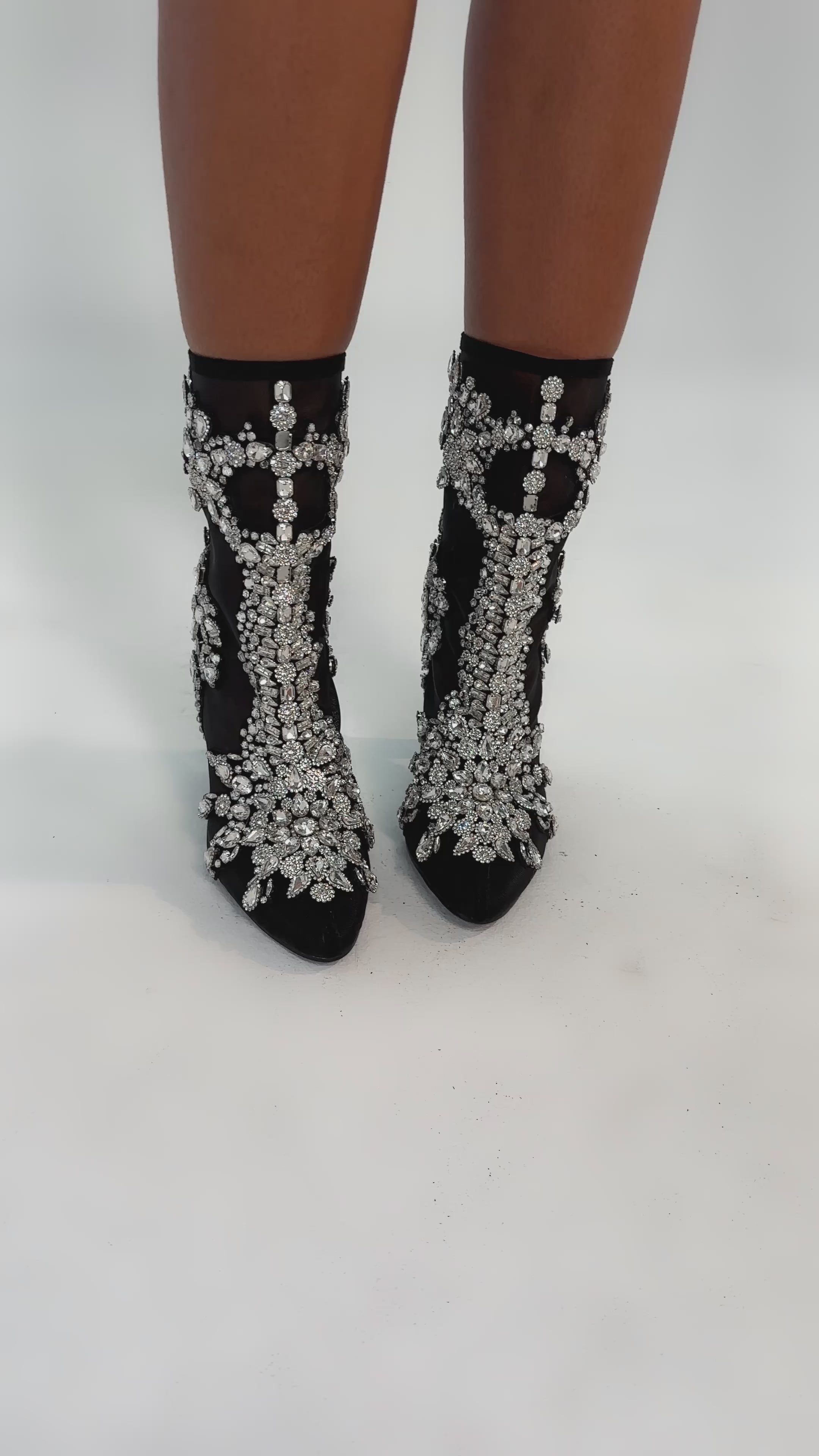 Video of woman modeling the AKIRA GOLD GRANDEUR BLACK EMBELLISHED COCKTAIL BOOTIE, featuring intricate rhinestone detailing and thick high heels, standing on a white background.