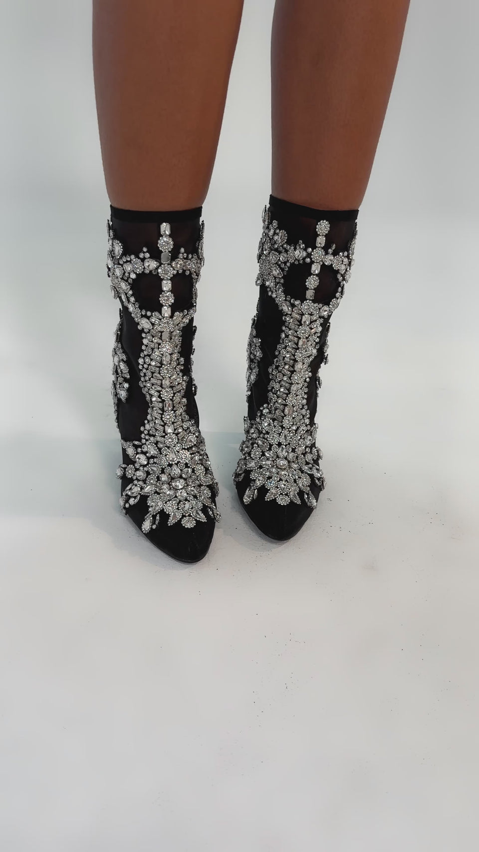 Video of woman modeling the AKIRA GOLD GRANDEUR BLACK EMBELLISHED COCKTAIL BOOTIE, featuring intricate rhinestone detailing and thick high heels, standing on a white background.