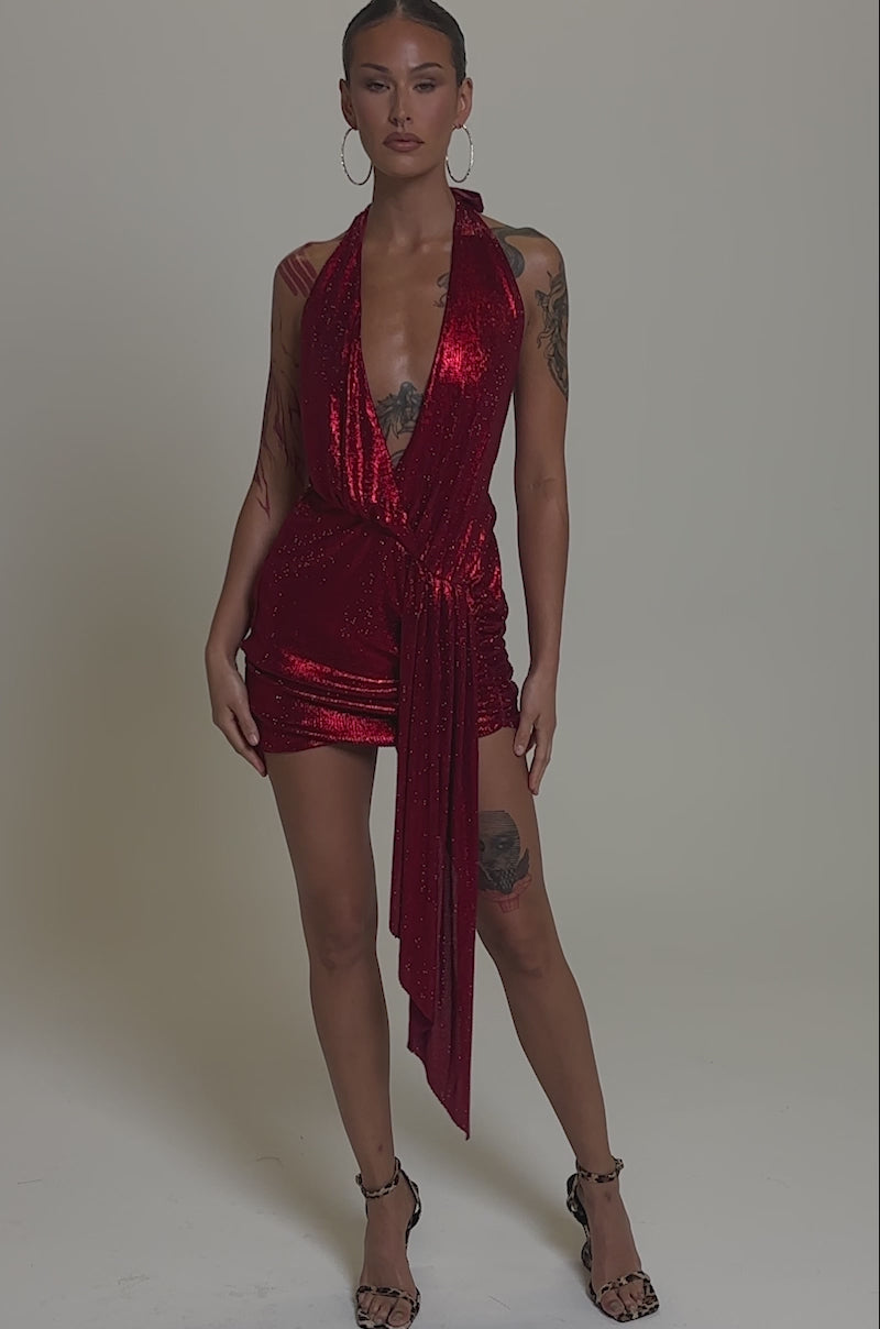 Video of woman modeling the DREAM GIRL RHINESTONE DRESS IN RED—a sparkly halter-neck mini with a draped asymmetrical sash—paired with hoop earrings, visible chest and arm tattoos, and slicked-back hair.