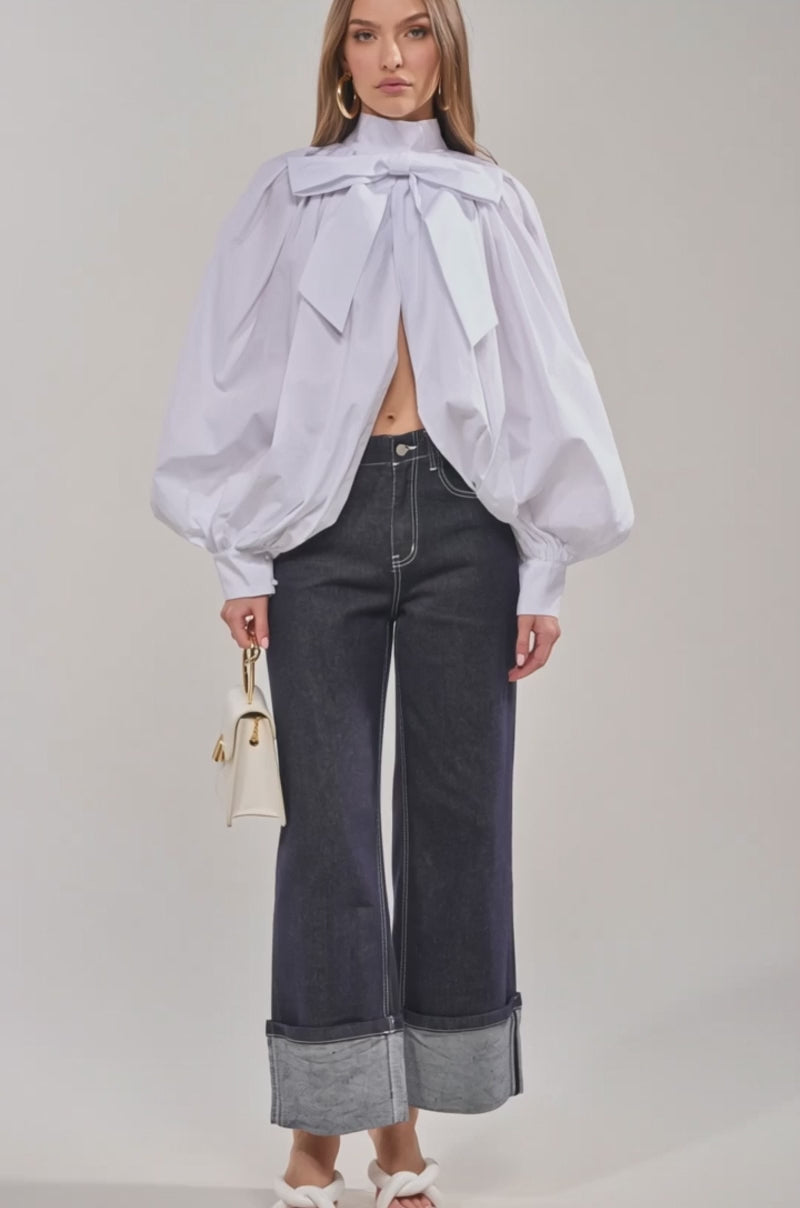 Video of woman modeling the BLACK TIE AFFAIR BLOUSE IN WHITE with voluminous sleeves, an oversized bow-tie neckline, and front cut-out, a woman pairs it with high-waisted dark jeans and holds a small handbag against a plain light background.