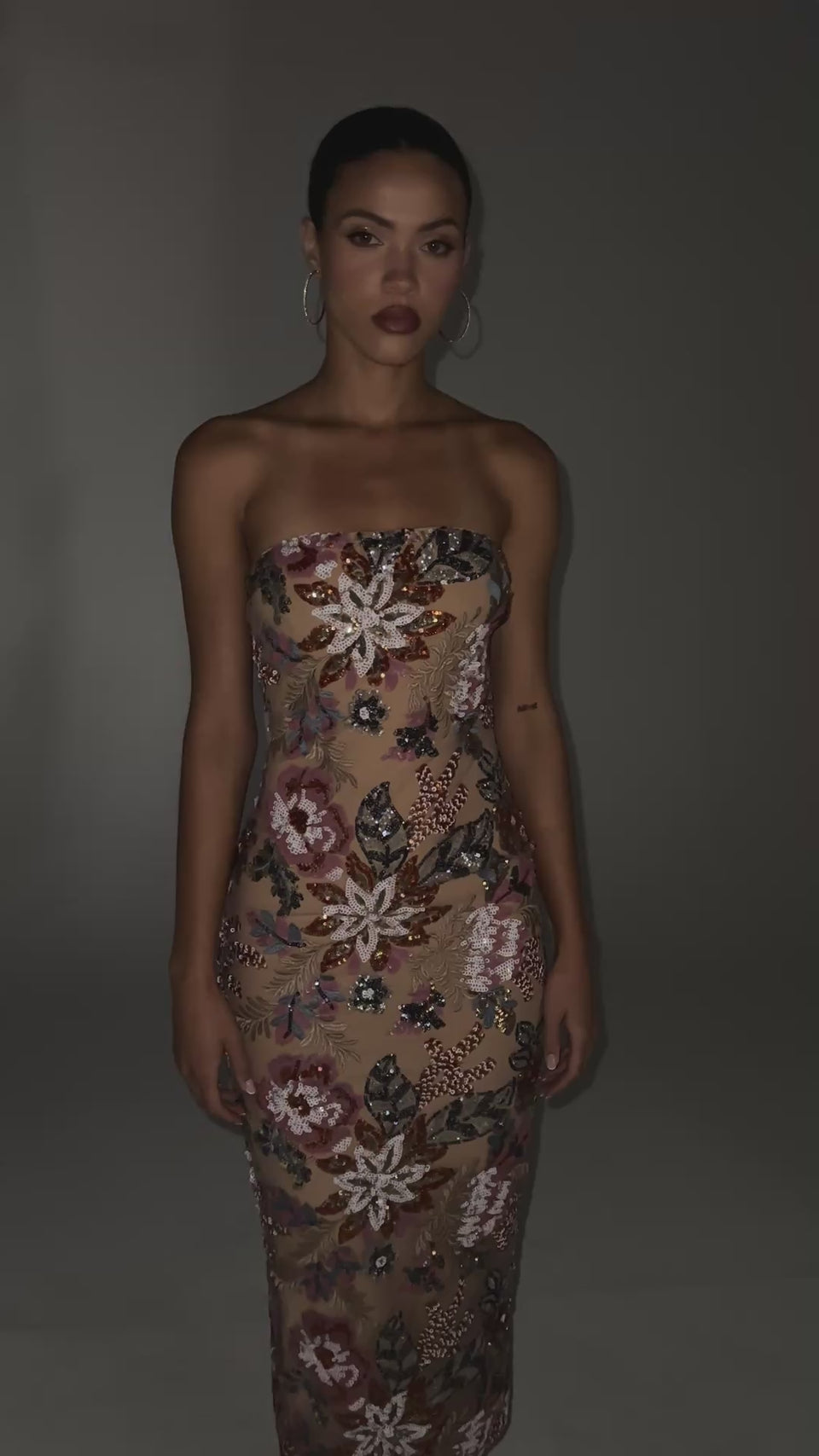 A video of a model wearing the SHOW ME LOVE EMBELLISHED MIDI DRESS.  Featuring a sleek body-contouring silhouette with a double-lined mesh base, a strapless neckline with inner elastic trim, intricate embroidered sequin floral detailing throughout for a richly textured, light-catching finish. Complete with a discreet back zip closure and a slit at the hemline for added movement.