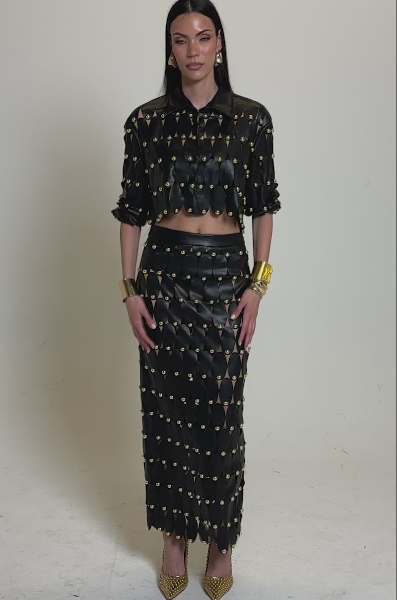 A video of a woman in a black long-sleeved crop top and THE SNEAK PEEK STUDDED PU MIDI SKIRT IN BLACK, both with gold circular accents. She accessorizes with gold bracelets, pointed heels, and has sleek hair against a neutral background.