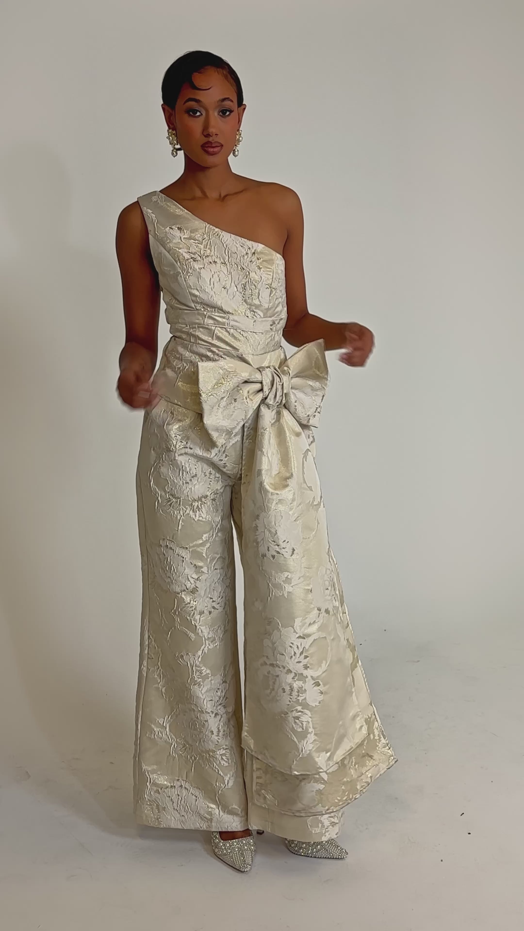 A video of a woman in the DRESSED TO IMPRESS TROUSER—a cream, one-shoulder design with intricate floral patterns, pearlescent jacquard, and a standout waist bow—set against a plain light background.