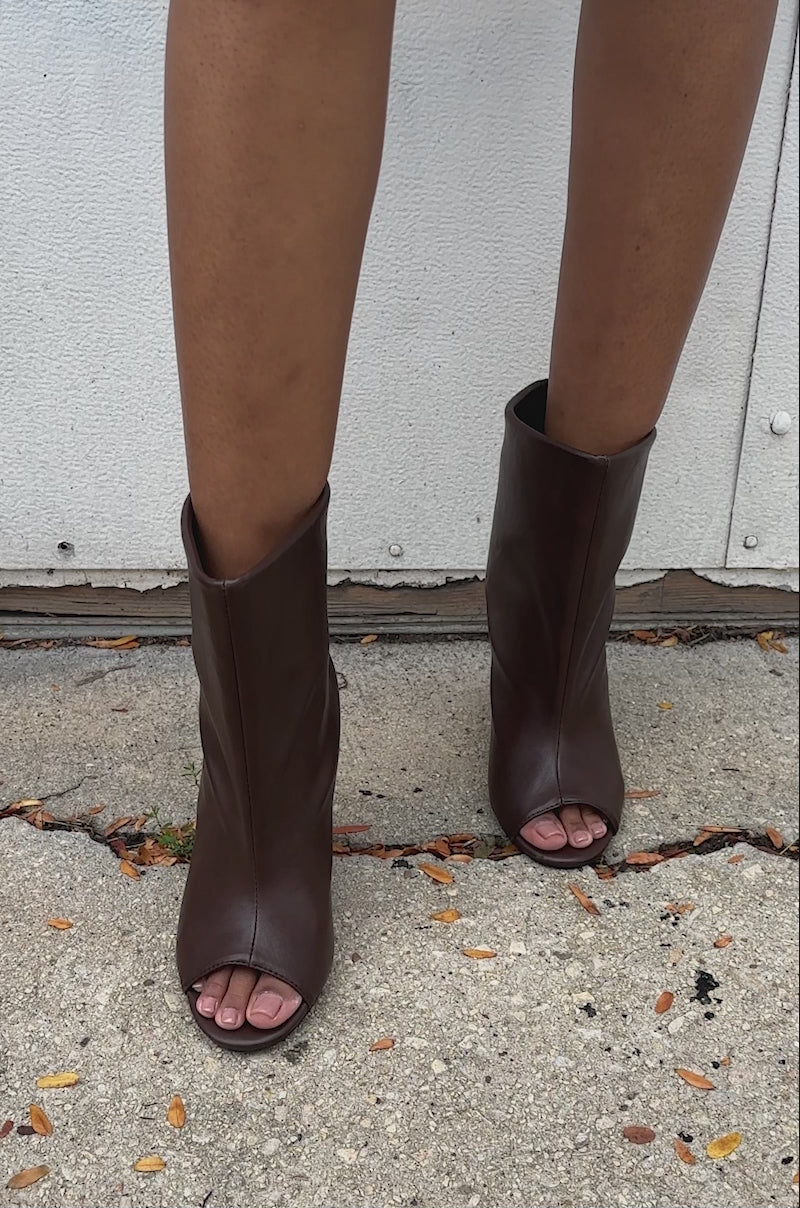 A video of a person with bare legs models the AZALEA WANG FAOI BROWN INVERTED WEDGE OPEN-TOE BOOTIE on a concrete surface against a white wall.