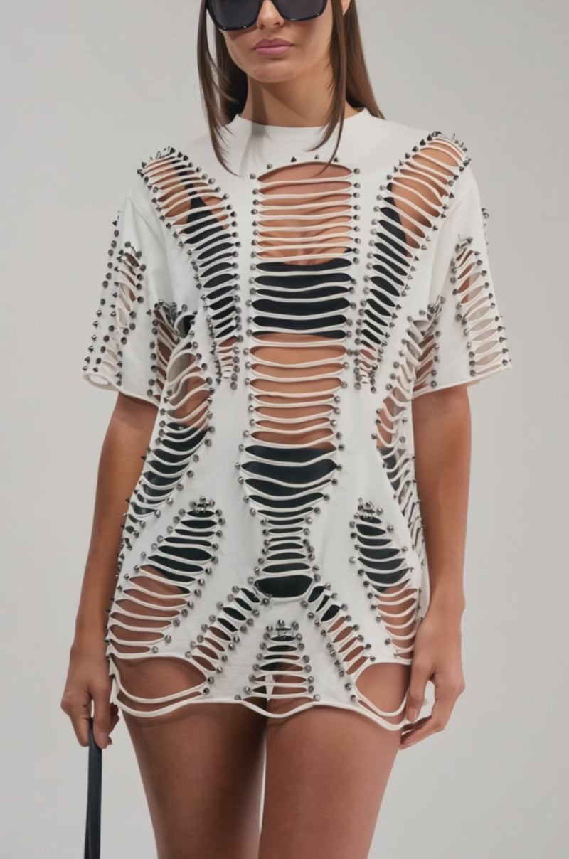 A video of a woman in the BARELY THERE DISTRESSED TEE—an oversized white top with large cutouts and stud embellishments—paired with bold black sunglasses against a plain background.