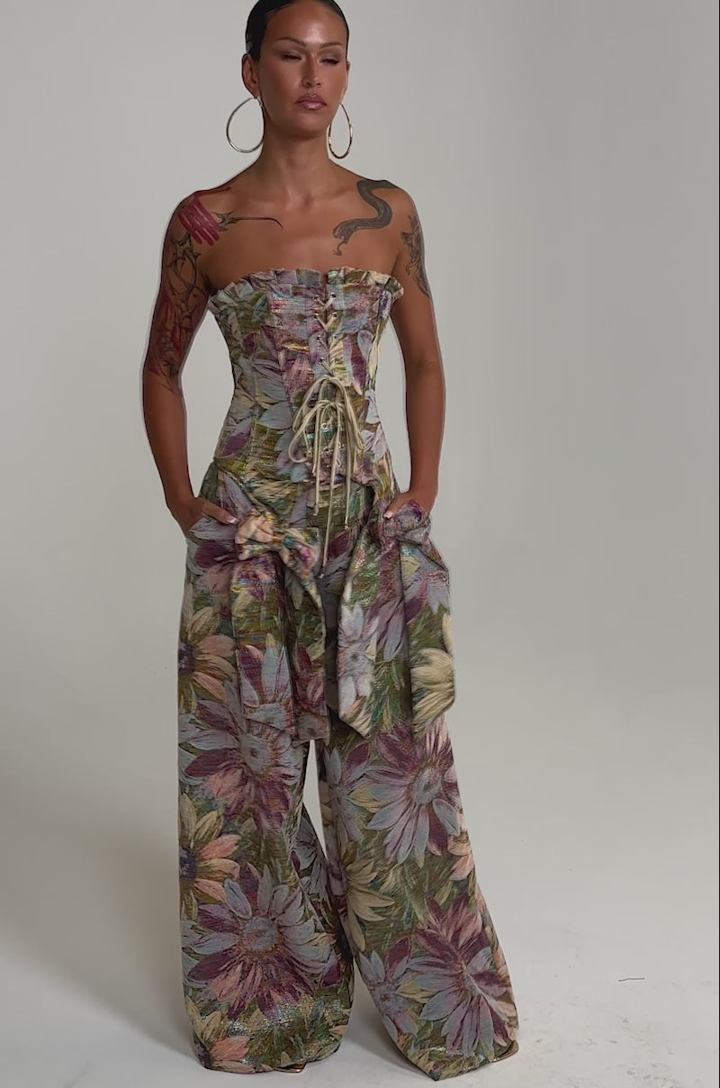 A video of a person wearing the SUNFLOWER FIELDS CORSET—a strapless, floral velvet top—paired with wide-leg metallic floral print pants, and hoop earrings.