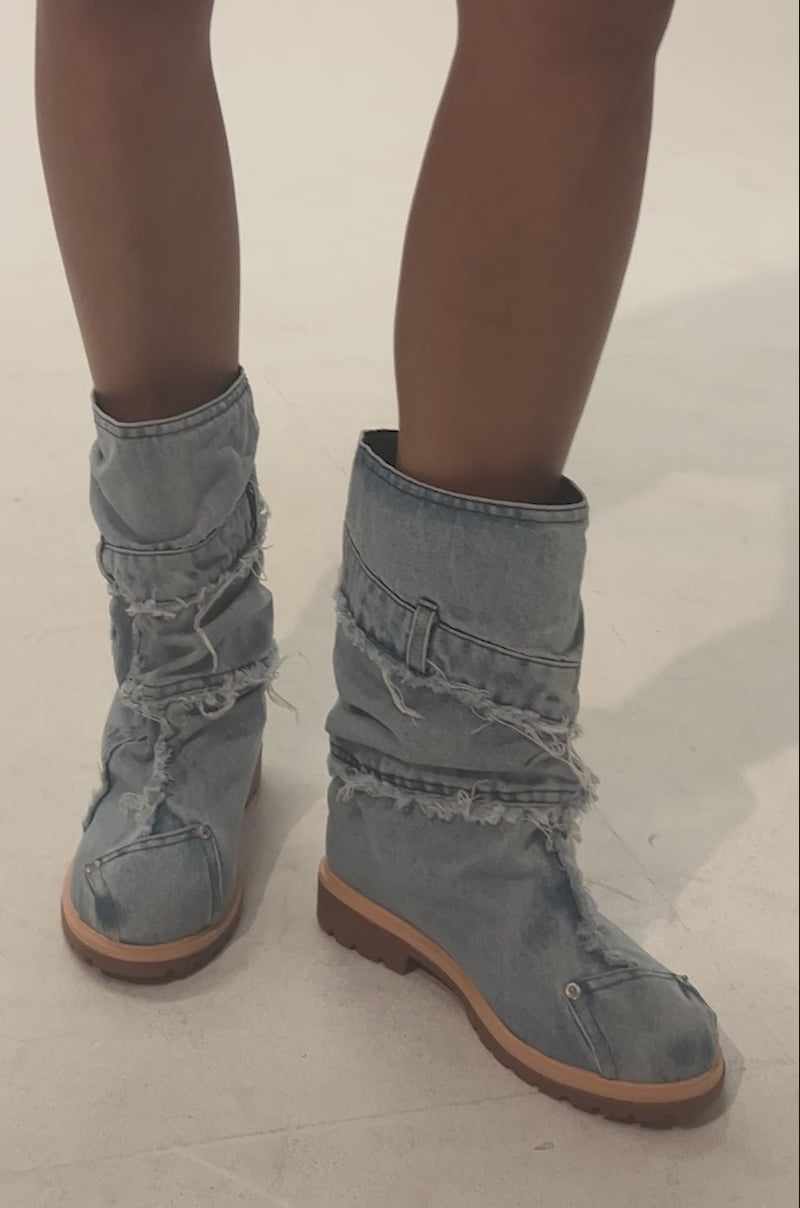 Video of woman modeling the AKIRA GOLD PAPILLION DENIM FLAT BOOTIE, a mid-calf boot with frayed edges and thick tan soles. Styled with bare legs, its rugged, distressed denim finish stands out against a plain background.