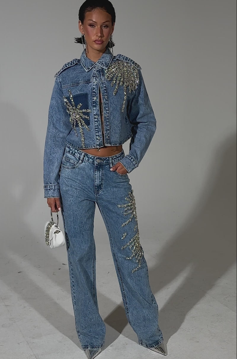 A video of a woman posing confidently in the SPARKLE AND SHINE QUEEN DENIM BUTTON DOWN, a cropped, rhinestone-embellished jacket paired with high-waisted jeans, a small white purse with silver accents, slicked-back hair, and pointed-toe shoes.