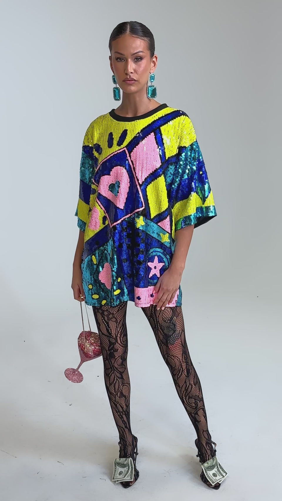 Video of woman modeling the QUEEN OF HEARTS SEQUIN TSHIRT DRESS, a bold, colorful oversized dress embellished with sequins, geometric shapes, hearts, and stars. She pairs it with blue earrings and slicked-back hair for a striking look.