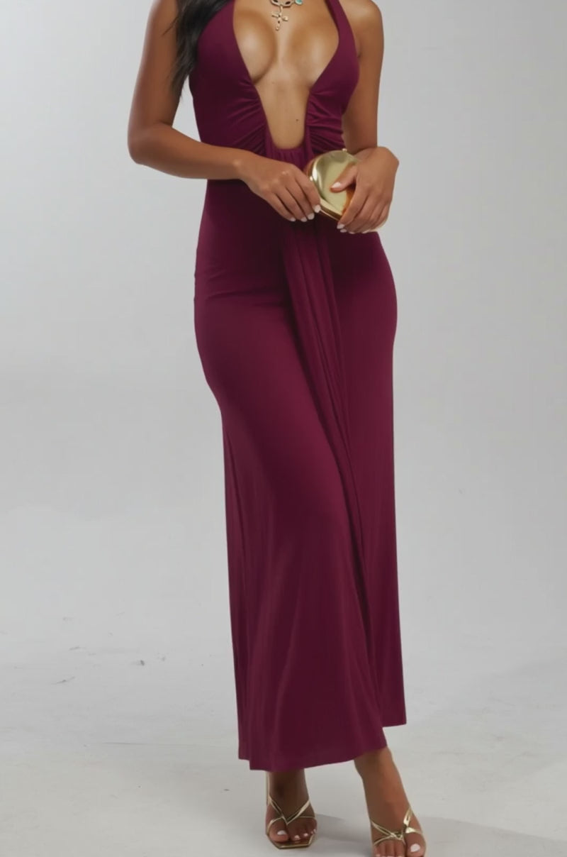 Video of woman modeling the MAKE YOU LOSE YOUR MIND MAXI DRESS IN BURGUNDY, paired with gold sandals, a statement necklace, and a gold clutch—an elegant vacation evening look against a plain light background.
