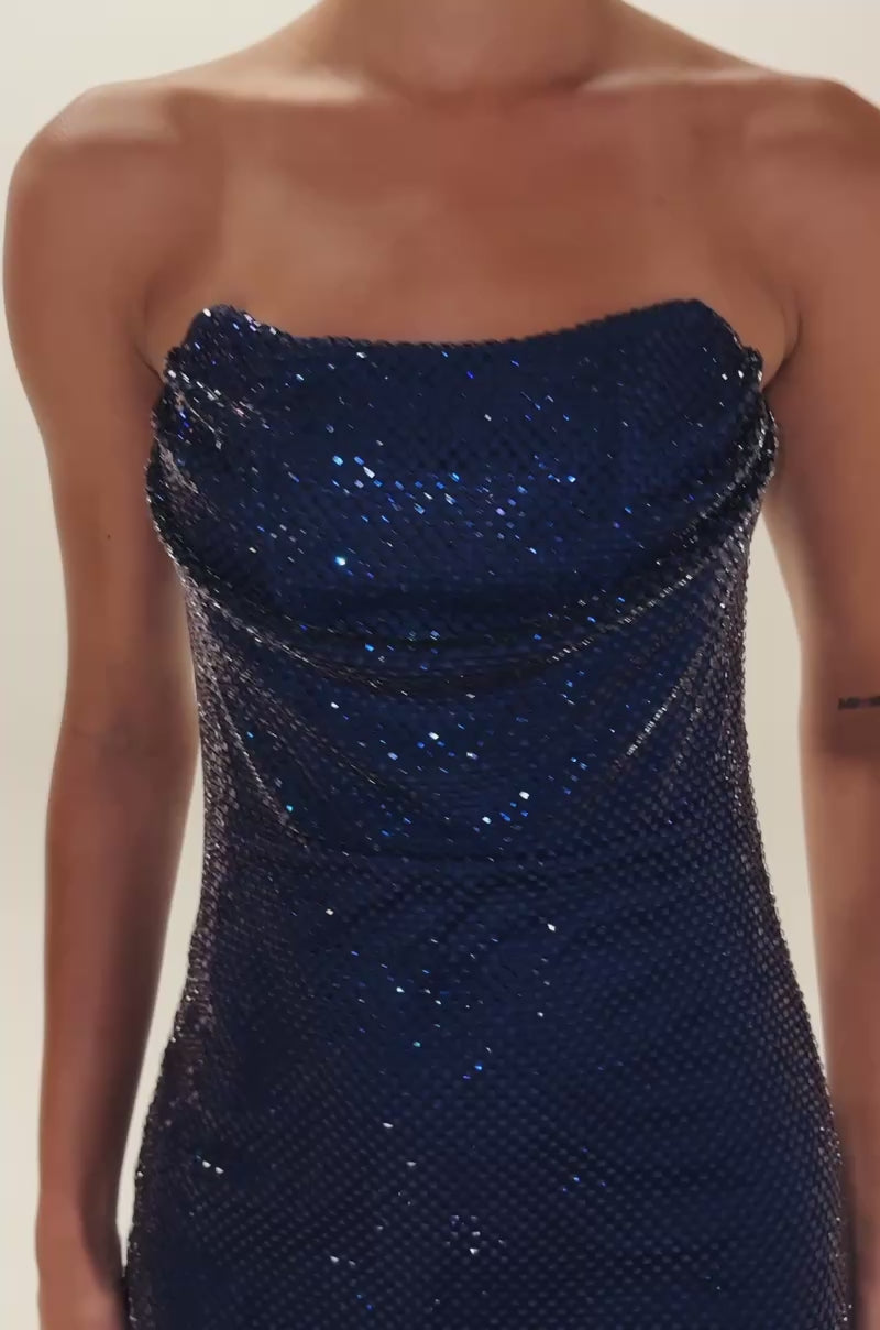Video of woman modeling the ELECTRIC AURA RHINESTONE STRAPLESS MINI DRESS, a shiny blue mesh-textured mini dress. She holds a small gold handbag, wears hoop earrings, and has slicked-back hair with a neutral expression.