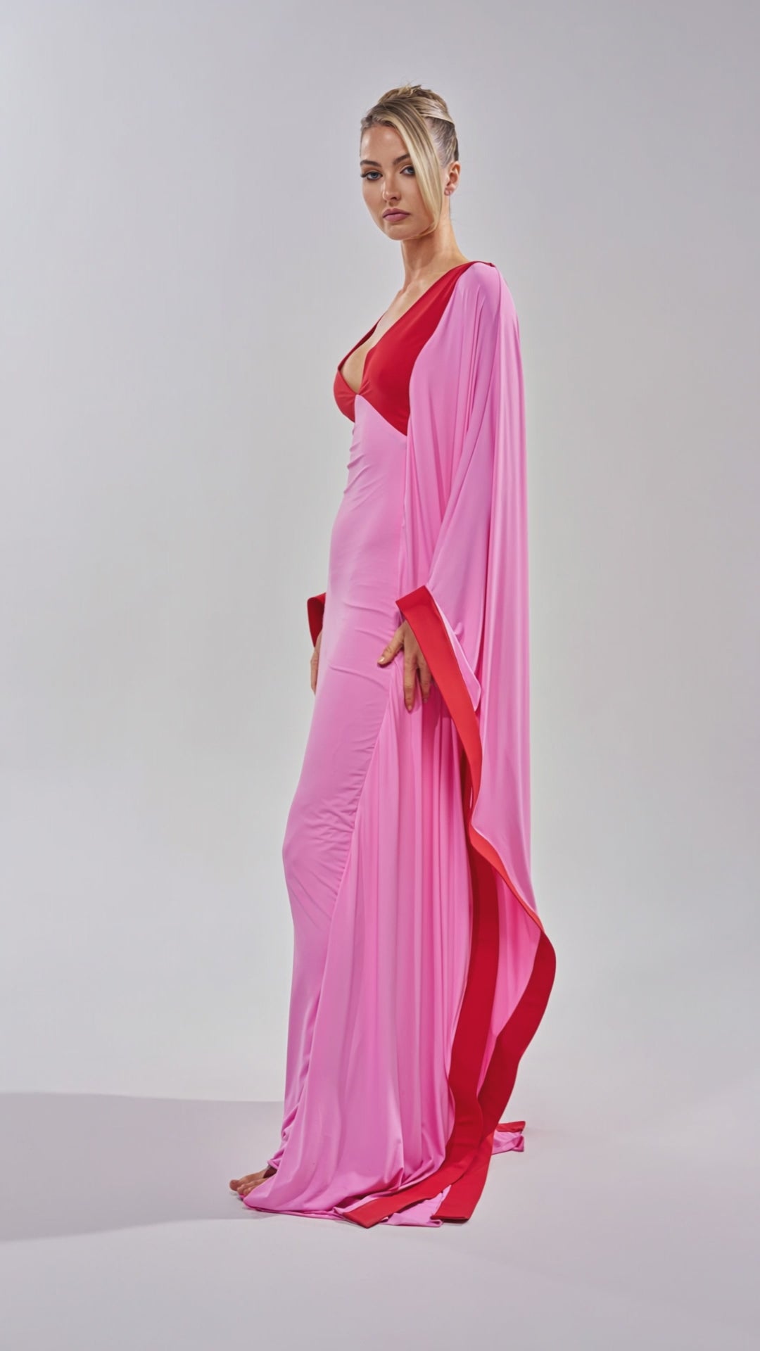 Video of woman modeling the GUEST OF HONOR BODYCON MAXI DRESS IN PINK—an elegant floor-length pink gown with a deep red neckline and red-trimmed, flowing sleeves