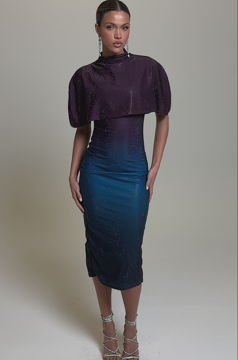 Video of woman modeling the CHIC AF RHINESTONE MIDI DRESS, a fitted sparkly design with a cape-like overlay that fades from deep purple to blue. She completes the look with strappy silver heels and long earrings against a plain background
